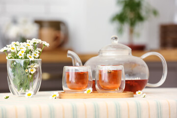 Wooden tray with teapot, cups of natural chamomile tea and flowers on table in kitchen