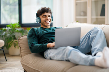 Smiling Arab Guy In Wireless Headphones Using Laptop At Home