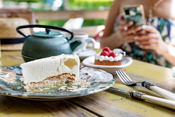 Cheesecake, strawberry tarts and tea on a table in a summer cafe. In the background is a girl who takes pictures of food on a smartphone.