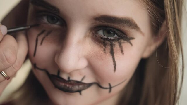 Mother making Halloween makeup to preteen girl daughter and painting spooky black lines around eyes closeup portrait. Pretty child with creepy cosmetics getting ready for holiday party