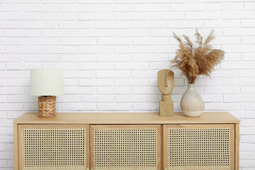 Wooden chest of drawers with lamp and pampas grass near light brick wall in room