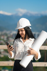 Beautiful young woman architect in a white helmet with a mobile phone and with a drawing in her hands on a construction site against the background of mountains. Construction concept. Vertical photo