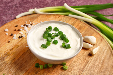 Board with bowl of tasty sour cream and sliced green onion on purple background, closeup