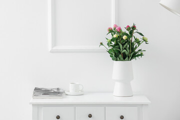 Vase with unopened peonies, cup of coffee and magazines on dresser near white wall