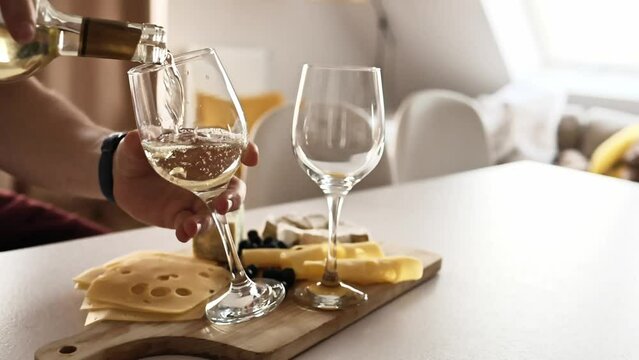 Man pouring white wine to glasses from bottle at kitchen with cheese plate on table. Transparent wineglasses and sommelier filling them with alcohol beverage