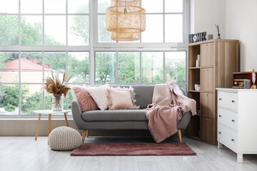 Interior of light living room with cozy grey sofa, shelving unit and chest of drawers near big window