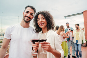 On foreground a multiracial couple toasting the wine glasses, smiling and looking at camera together, at background their friends clinking on rooftoop a party celebration. Patio social reunion at home