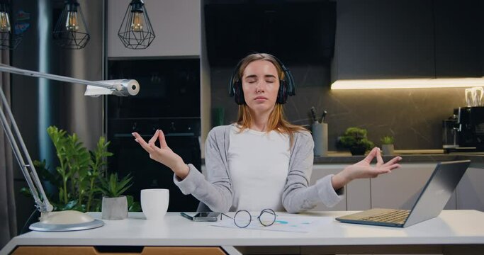 Pretty Girl In Headphones Sitting The Workplace Work On Laptop Computer, Meditating, Doing Yoga Breathing Exercise With Namaste Hands Position At Home Office. Calm Serene Freelancer Woman Taking Break