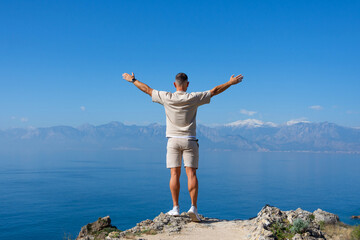 Man stands with arms raised on rock blue ocean mountain peaks horizon. Active holiday adventure,...