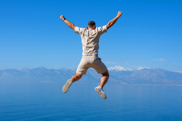 Young man jumping on rock with his hands up blue ocean mountain peaks horizon. Active holiday...