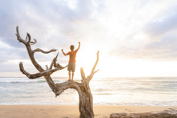 Horizontal of a man enjoying a beautiful sunset standing with his arms raised on a dry tree trunk in the middle of the beach. 