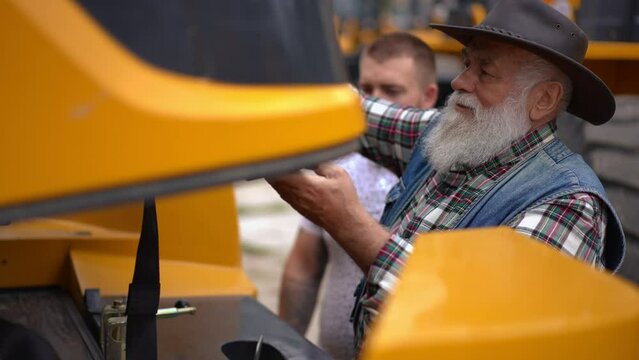 Senior man opening front part of huge tractor talking with young guy outdoors. Experienced professional Caucasian farmer choosing new vehicle with colleague. Slow motion