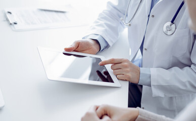 Doctor and patient sitting at the table in clinic. The focus is on female physician's hands using tablet computer, close up. Medicine and healthcare concept