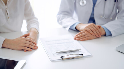 Fototapeta premium Doctor and patient discussing current health questions while sitting near of each other and using clipboard at the table in clinic, just hands closeup. Medicine concept