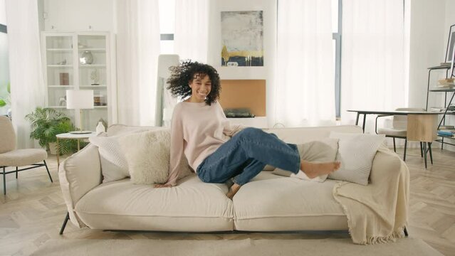 Cheerful African American positive woman jumping alone barefoot on sofa in modern living room at home. Happy female celebrate moving relocation day enjoy new rented apartment. Stress-free fun weekend