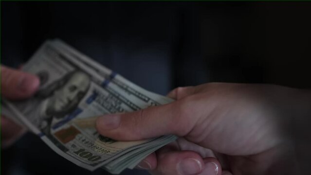 Two people are trying to divide money among themselves in a dark room, man's hand is pulling pack of 100 dollar bills strongly, close up, black vignette.