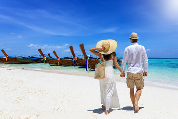 A tourist couple walks down a beautiful beach in Krabi, Thailand with fine sand a longtail boats...