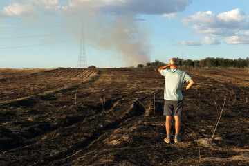 A man with his back turned looks concerned at his burnt field after the fire. He is placed to the right of the image.