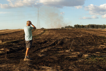 Man on his back looks at his burned field after fire. He has his hands on his head. There is still...