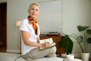 Good-looking proud confident and successful 50-years-old businesswoman sitting on sofa in her living-room, writing in her diary or notepad, making notes, or noting down new business plan ideas