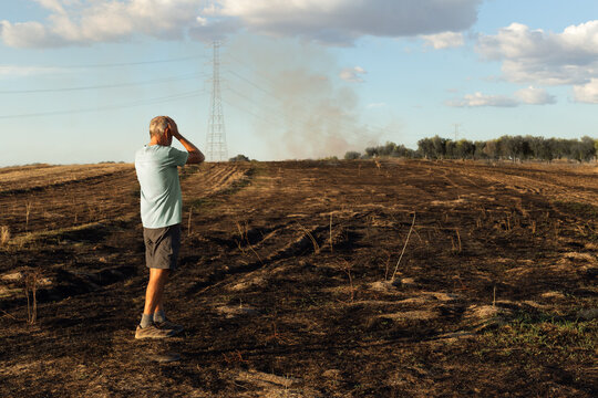 Man on his back looks at his burned field after fire. He has his hands on his head. There is still smoke from bottom. He is placed to the left of the image.