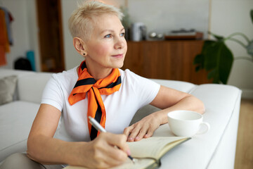 Dreamy pensive elderly female novelist sitting on couch in living-room leaning on its back and...