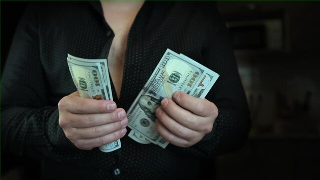 Man in black unbuttoned shirt fast counts dollar bills and puts them in pack holding them in front of him, on dark background, close up. Concept of financial wealth, money, and power.