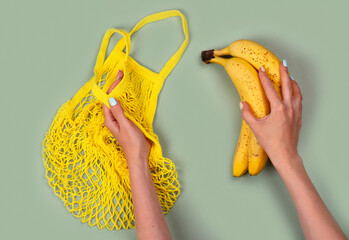 Yellow mesh string bag and female's hands holding bunch of bananas on green. International plastic free day background. 3rd of July. Copy space