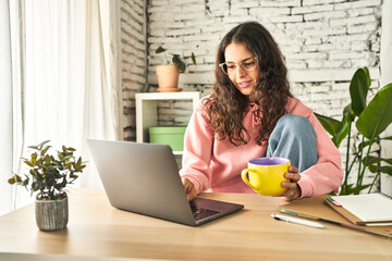 A young woman, focused and productive, working at her home desk with a laptop, sipping coffee, and feeling refreshed.