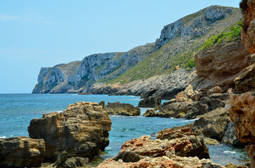 Rocks in the water on the Mediterranean coast of Spain overlooking the cliffs and mountains on the coast and the ocean under blue sunny sky