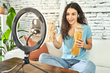 A young Caucasian influencer, setting up for a video recording on her sofa with a ring light and...