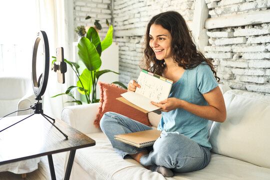 A young Caucasian influencer, sharing book tips and reviews, capturing creative videos on her sofa with a ring light and mobile device.