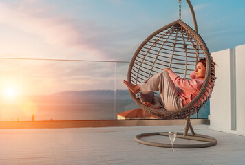 Beautiful middle-aged barefoot woman lying in comfortable hanging chair on open house terrace and...