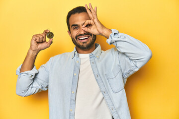 Young Latino man holding a Bitcoin coin, yellow studio background, excited keeping ok gesture on eye.