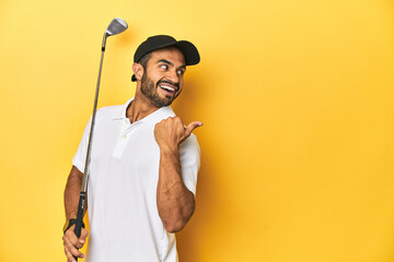 Young Latino golfer with club and cap on a yellow studio background, points with thumb finger away,...
