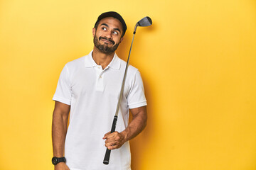 Young Latino golfer with club and cap on a yellow studio background, dreaming of achieving goals and purposes