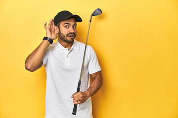 Young Latino golfer with club and cap on a yellow studio background, trying to listening a gossip.