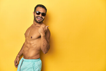 Fit young Latino man in swimwear and sunglasses, yellow studio background, points with thumb finger away, laughing and carefree.