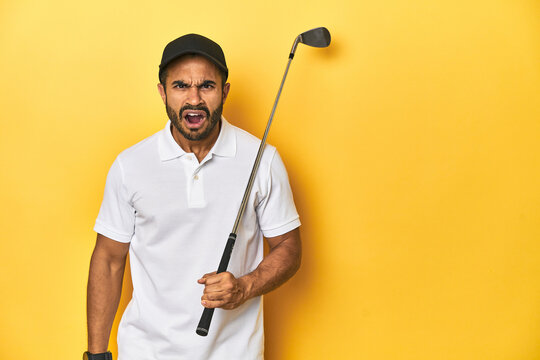 Young Latino golfer with club and cap on a yellow studio background, screaming very angry and aggressive.