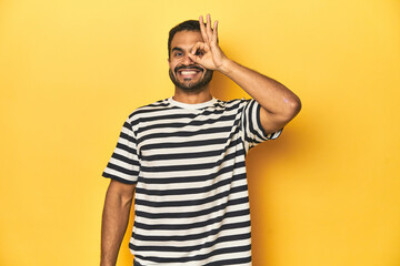 Casual young Latino man against a vibrant yellow studio background, excited keeping ok gesture on...