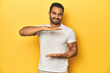 Casual young Latino man against a vibrant yellow studio background, holding something with both hands, product presentation.