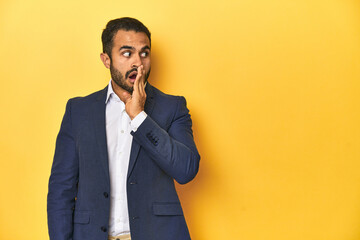 Professional young Latino man in business suit, yellow studio background, is saying a secret hot braking news and looking aside