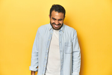Casual young Latino man against a vibrant yellow studio background, laughs and closes eyes, feels relaxed and happy.