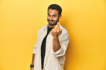Casual young Latino man against a vibrant yellow studio background, pointing with finger at you as if inviting come closer.