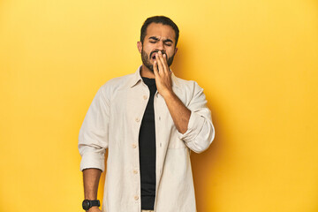 Casual young Latino man against a vibrant yellow studio background, yawning showing a tired gesture...