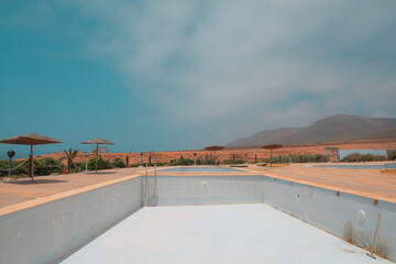 Large empty hotel swimming pool without water in Legzira, Morocco. Blue day sky, huts, mountain...