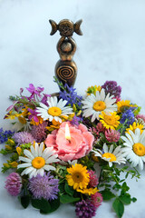 Triple Goddess figurine with candle and colorful flowers close up, abstract light background. Symbol of the Triune Moon. esoteric ritual for Litha, Midsummer. witchcraft, wiccan spiritual practice