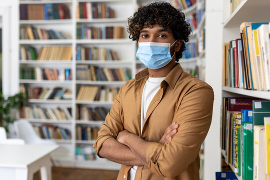 Portrait of young student inside library, man with crossed arms looking at camera, hispanic man with protective face mask visiting academic library and bookstore.