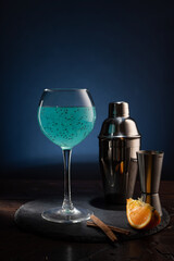 Bar composition on the table. Wine glass with a bright turquoise drink and an iron shaker. Orange slices and cinnamon sticks.