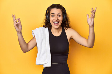 Athletic woman holding a towel, yellow background, receiving a pleasant surprise, excited and...